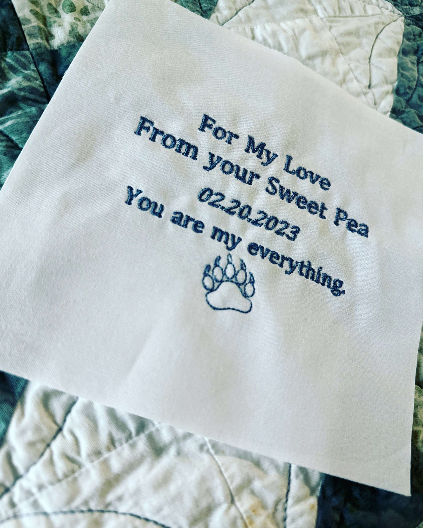 Embroidered quilt label with personalized message and paw print graphic on white cotton fabric.