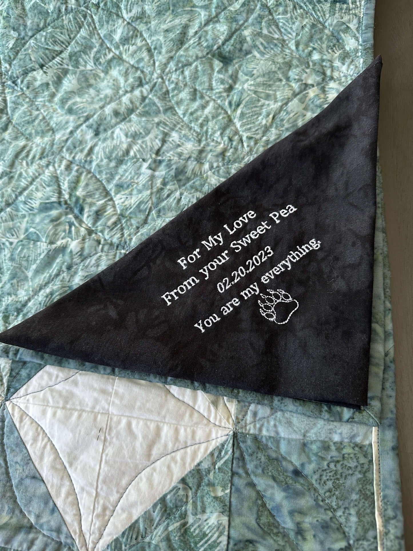 Personalized embroidered quilt label with custom message and paw graphic on 100% quilt cotton, unfinished edges visible.