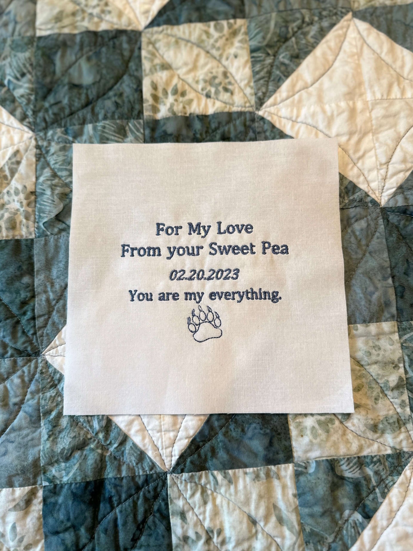 Embroidered quilt label on cotton with personalized text and paw print graphic, displayed on a quilt background.