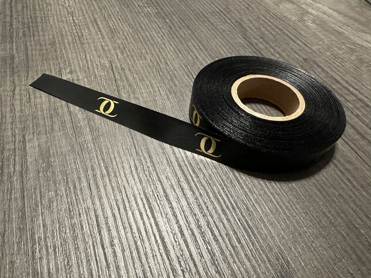 Roll of black satin ribbon with gold logo on wooden surface, customizable for sewing projects.