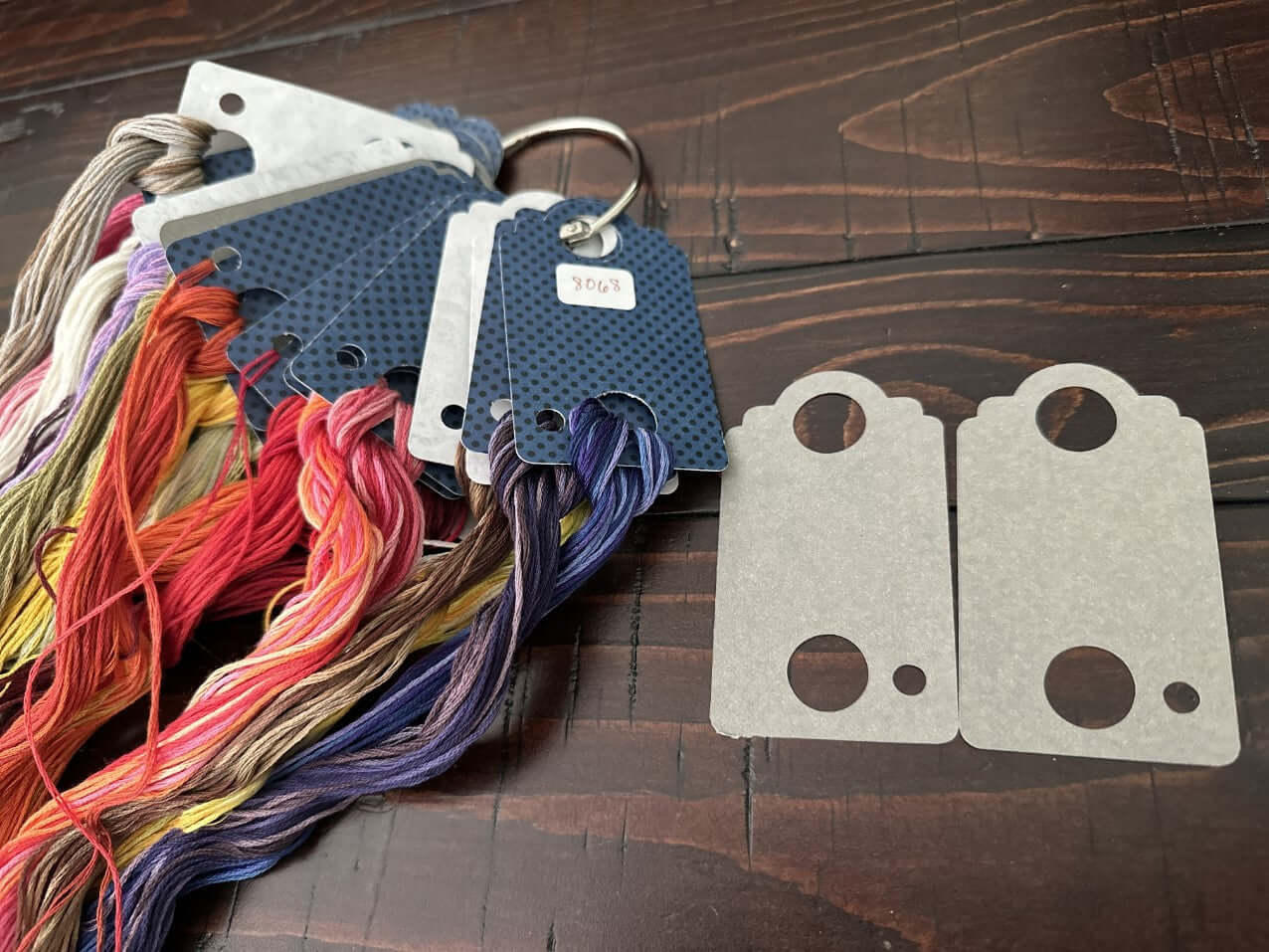 Colorful embroidery floss on thread drop holders with two unpainted floss holder templates on a wooden surface.