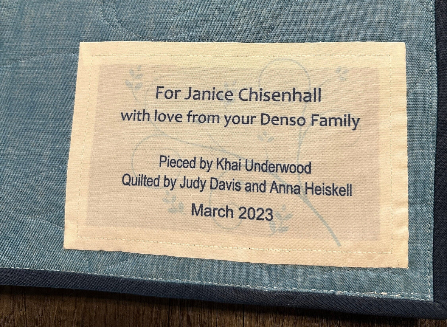 Quilt label with personalized text: 'For Janice Chisenhall, with love from your Denso Family. Pieced by Khai Underwood, Quilted by Judy Davis and Anna Heiskell, March 2023.'