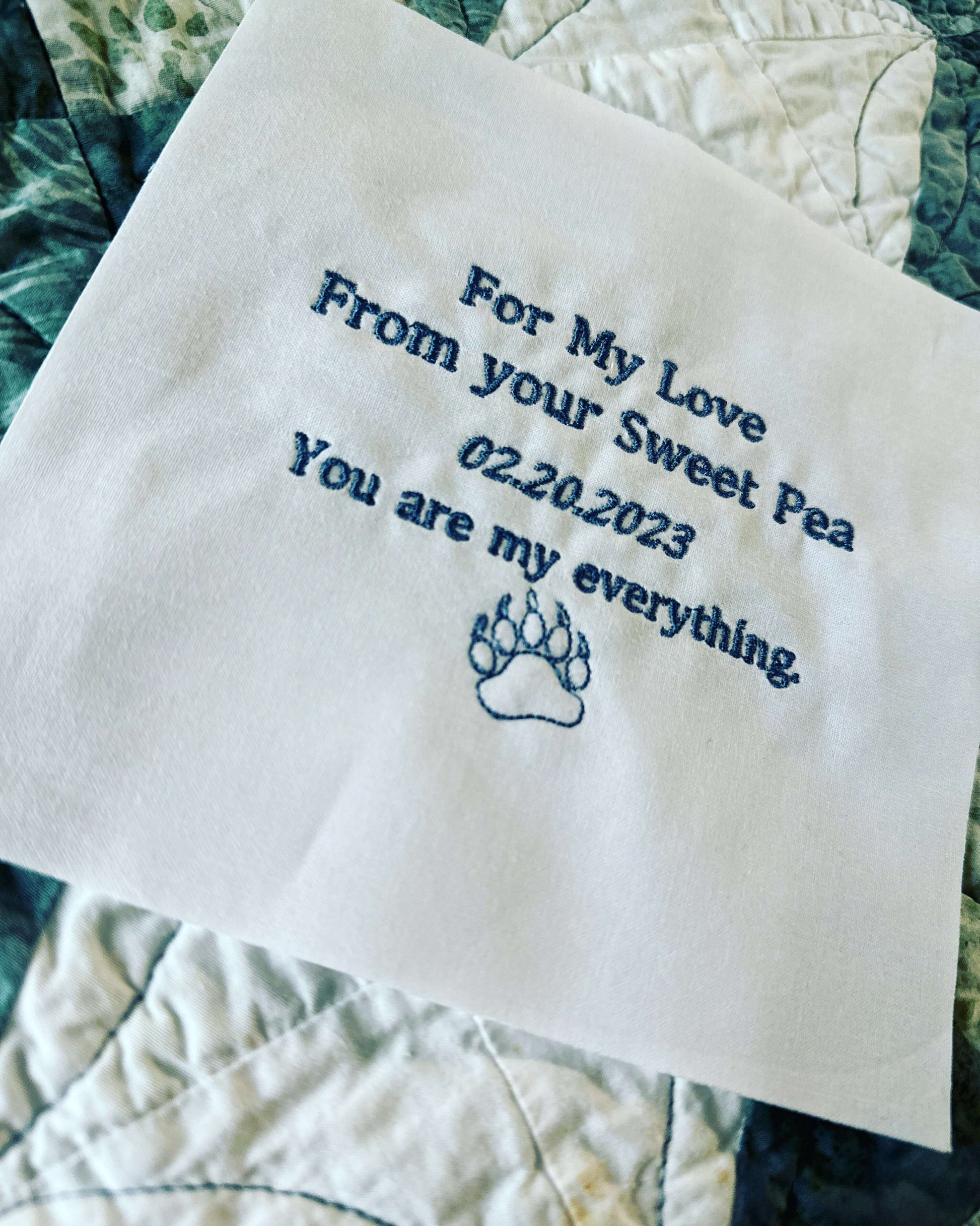 Embroidered quilt label with personalized message and paw print graphic on white cotton fabric.