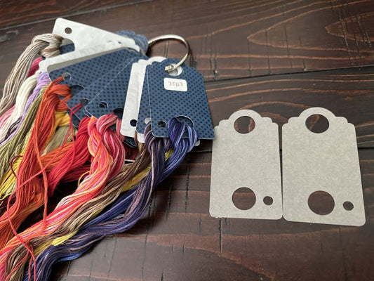 Colorful embroidery floss on thread drop holders with two unpainted floss holder templates on a wooden surface.